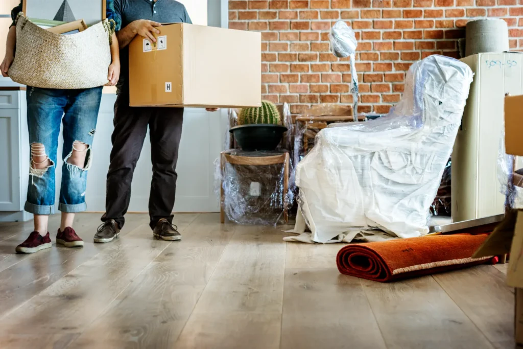 Zwei Personen mit Umzugskartons stehen in einem Raum mit Holzböden, eingepackten Möbeln und Zimmerpflanzen, was auf einen Umzug in einen neuen Raum hindeutet - perfekt für eine reinigungsfirma zürich, die dafür sorgt, dass alles makellos ist.